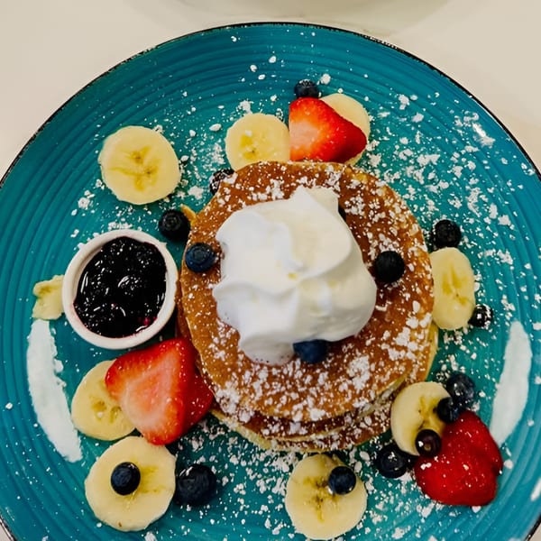 Pancakes with whipped cream, blueberries, strawberries, bananas, and a side of jam on a blue plate.
