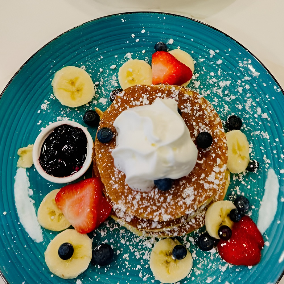 Pancakes with whipped cream, blueberries, strawberries, bananas, and a side of jam on a blue plate.
