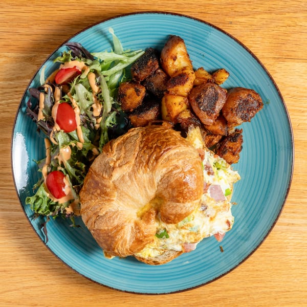 A croissant sandwich with omelet, side salad, and roasted potatoes on a blue plate.